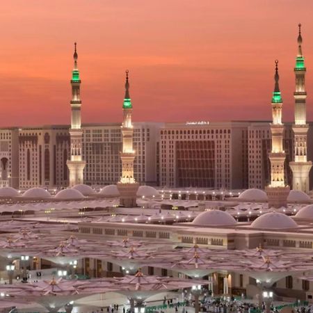 Hotel Intercontinental Dar Al Iman Madinah By Ihg Medína Exteriér fotografie The photo depicts a scenic view of a mosque complex, likely during sunset, with several minarets illuminated and showcasing green lights. The architecture features domed roofs, and the setting includes a backdrop of buildings, all bathed in a warm, o