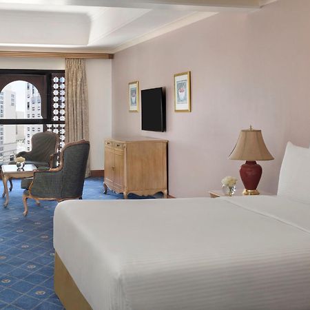 Hotel Intercontinental Dar Al Iman Madinah By Ihg Medína Exteriér fotografie The photo shows a well-furnished hotel room. It features a large bed with a white duvet, two bedside lamps, and decorative paintings on the walls. There is a comfortable seating area with two chairs and a small table. A wooden dresser is positioned a