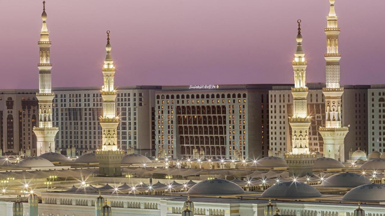 Hotel Intercontinental Dar Al Iman Madinah By Ihg Medína Exteriér fotografie The photo depicts an architectural scene, likely from a mosque complex, featuring prominent minarets that are illuminated. The setting appears to be during twilight, with a soft gradient in the sky. In the background, a large building is visible, pos
