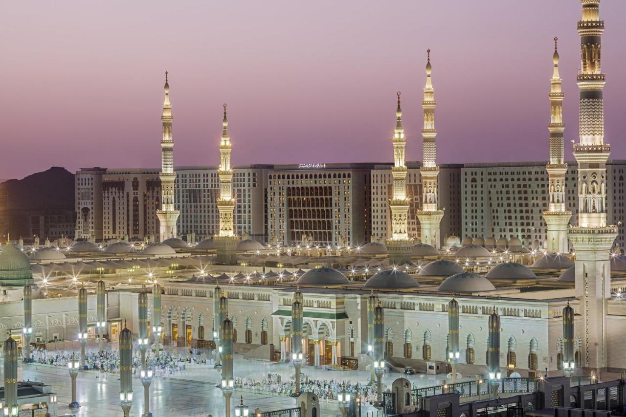 Hotel Intercontinental Dar Al Iman Madinah By Ihg Medína Exteriér fotografie The photo shows a view of an expansive mosque complex, featuring several tall minarets that are illuminated against a twilight sky. The structure appears grand and is likely adorned with intricate architectural details. There are people gathered in t