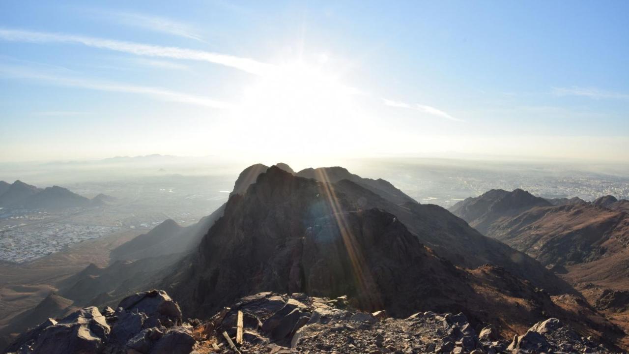 Hotel Intercontinental Dar Al Iman Madinah By Ihg Medína Exteriér fotografie The photo shows a scenic view from a mountain top, with peaks silhouetted against the bright sun. The sun is positioned low in the sky, creating a warm glow and illuminating the landscape below. In the foreground, there are rocky terrains, while the 