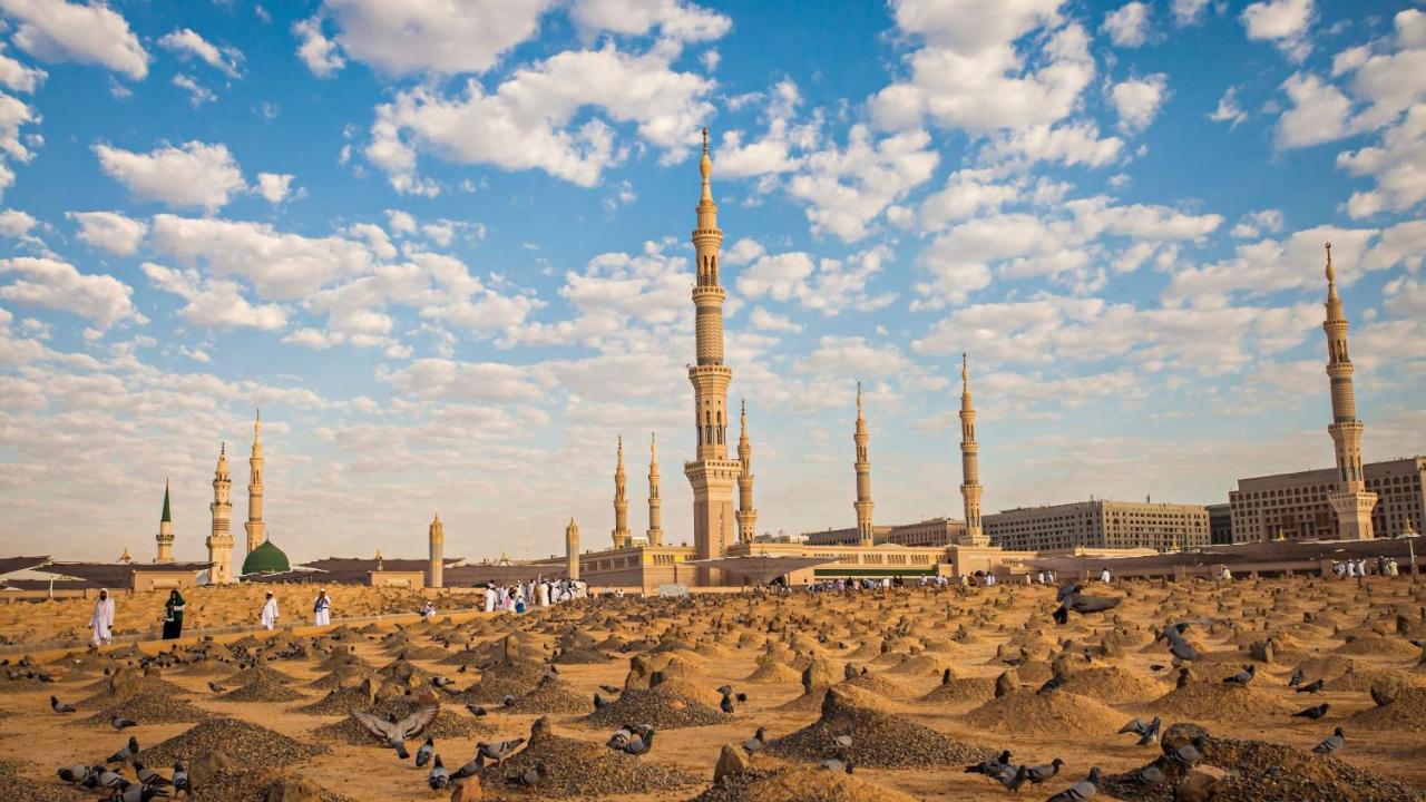 Hotel Intercontinental Dar Al Iman Madinah By Ihg Medína Exteriér fotografie The photo shows a landscape that features a large mosque with a tall minaret surrounded by smaller minarets. In the foreground, there are numerous grave markers arranged in a desert-like setting. The sky is bright with scattered clouds, and there are