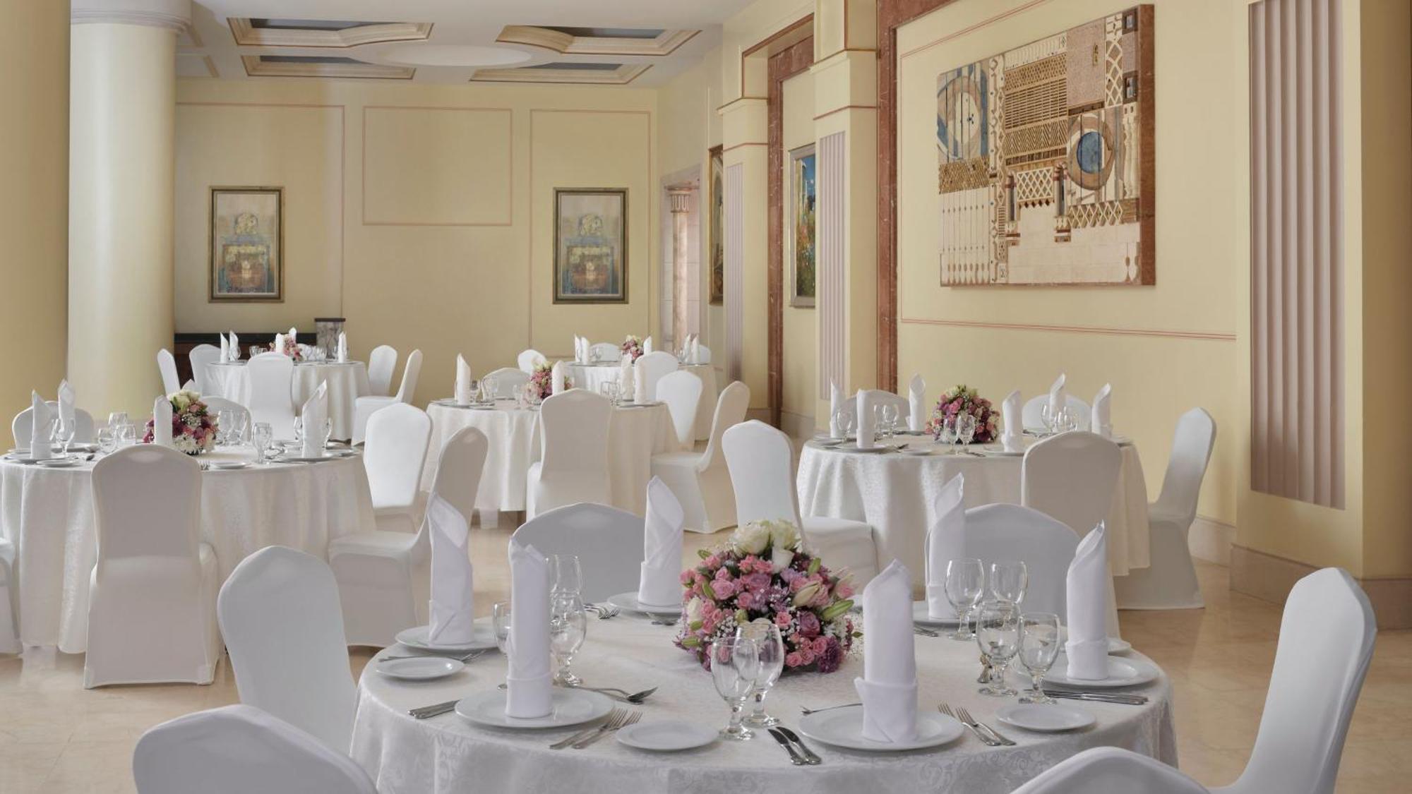 Hotel Intercontinental Dar Al Iman Madinah By Ihg Medína Exteriér fotografie The photo shows an elegantly arranged dining area, likely set up for a formal event or reception. There are several round tables covered with white tablecloths, each adorned with floral centerpieces featuring pink flowers. The tables are set with pla