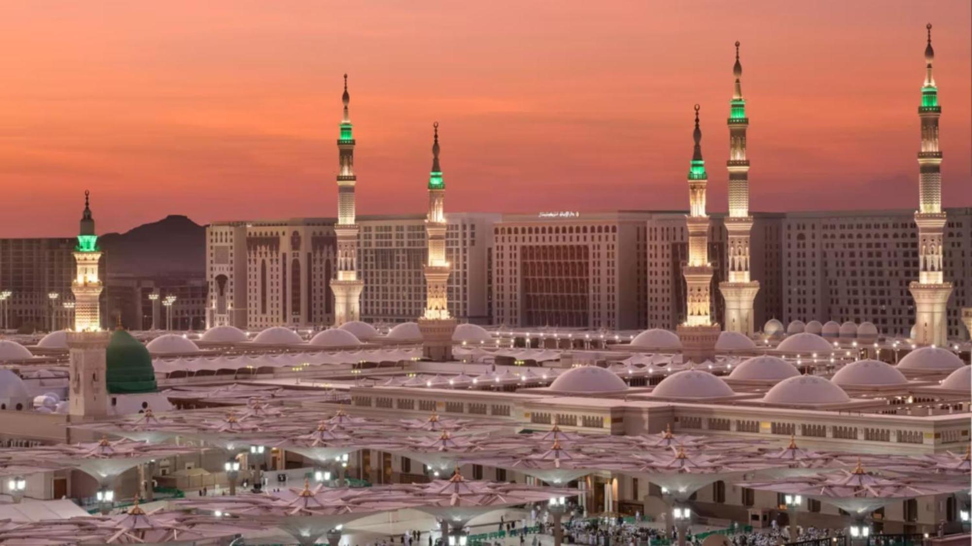 Hotel Intercontinental Dar Al Iman Madinah By Ihg Medína Exteriér fotografie The photo depicts a scenic view of a mosque complex, likely during sunset, with several minarets illuminated and showcasing green lights. The architecture features domed roofs, and the setting includes a backdrop of buildings, all bathed in a warm, o