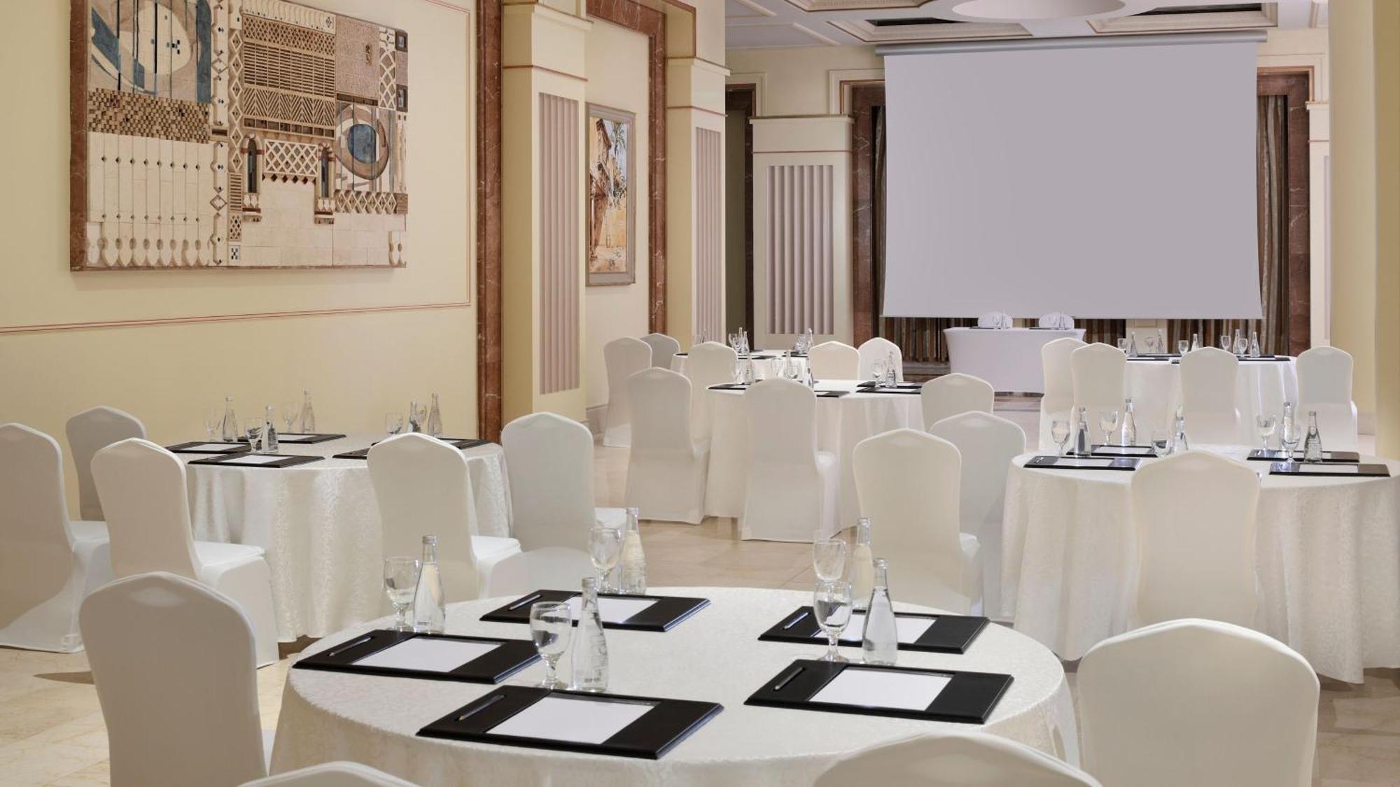 Hotel Intercontinental Dar Al Iman Madinah By Ihg Medína Exteriér fotografie The photo depicts an elegantly arranged event space. There are several round tables covered with white tablecloths and surrounded by white chairs. Each table is set with a black folder or menu and a glass of water. The walls are adorned with artworks