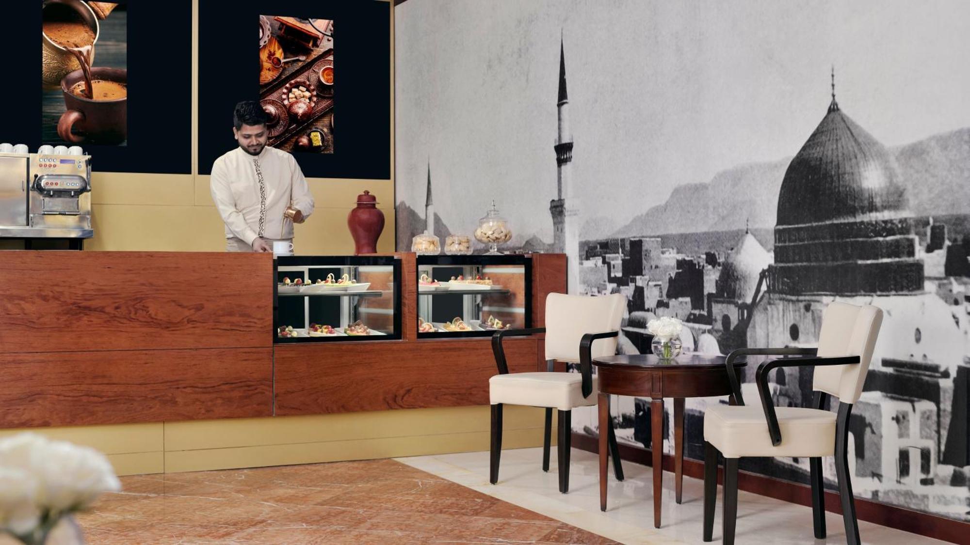 Hotel Intercontinental Dar Al Iman Madinah By Ihg Medína Exteriér fotografie The photo shows an elegant cafe or dessert shop interior. A man in traditional attire is standing behind a wooden counter, where various pastries and sweets are displayed. The walls feature a large black-and-white mural of a cityscape, possibly depic