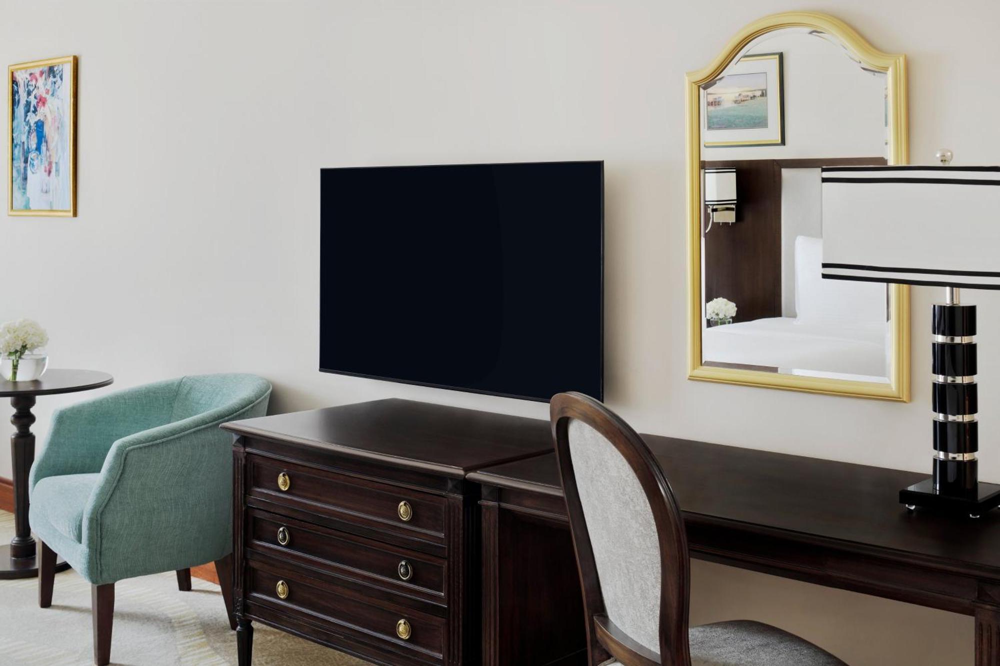 Hotel Intercontinental Dar Al Iman Madinah By Ihg Medína Exteriér fotografie The photo shows a hotel room interior featuring a mounted flat-screen television on the wall. Below the TV, there is a dark wooden dresser with drawers. To the right, there's a wooden desk and a chair with a light-colored seat. A small table with a l