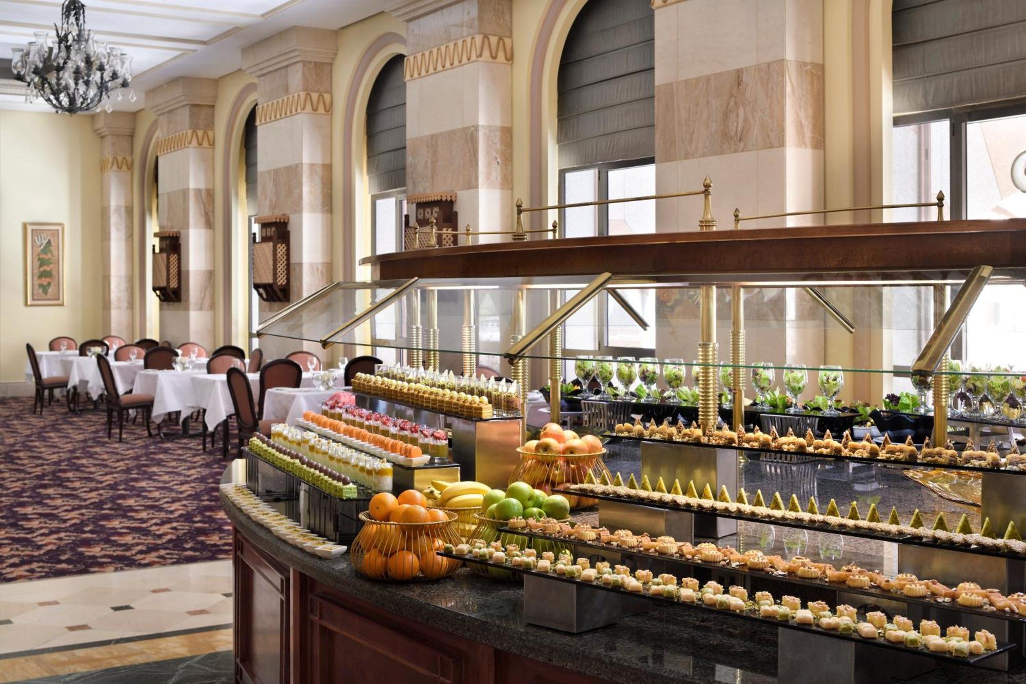 Hotel Intercontinental Dar Al Iman Madinah By Ihg Medína Exteriér fotografie The photo depicts an elegant dining area featuring a buffet-style display. In the foreground, a beautifully arranged buffet counter showcases an array of desserts, including miniature pastries and colorful fruit. The setting has a sophisticated ambia