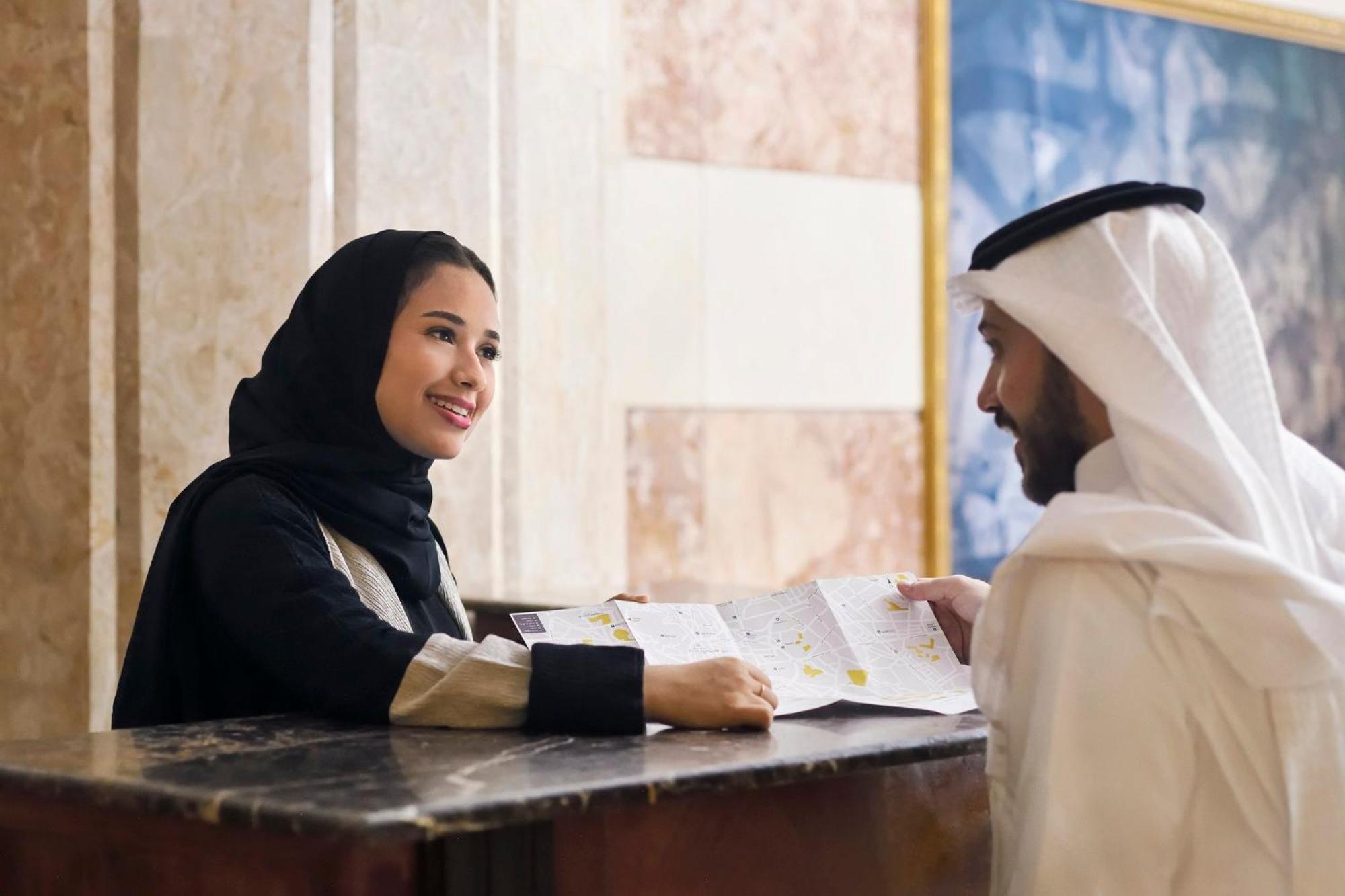 Hotel Intercontinental Dar Al Iman Madinah By Ihg Medína Exteriér fotografie The photo depicts a woman in a black hijab and a light-colored outfit smiling as she interacts with a man wearing traditional Emirati attire, including a white kandura and ghutrah (headscarf). They are at a reception desk in what appears to be a hote