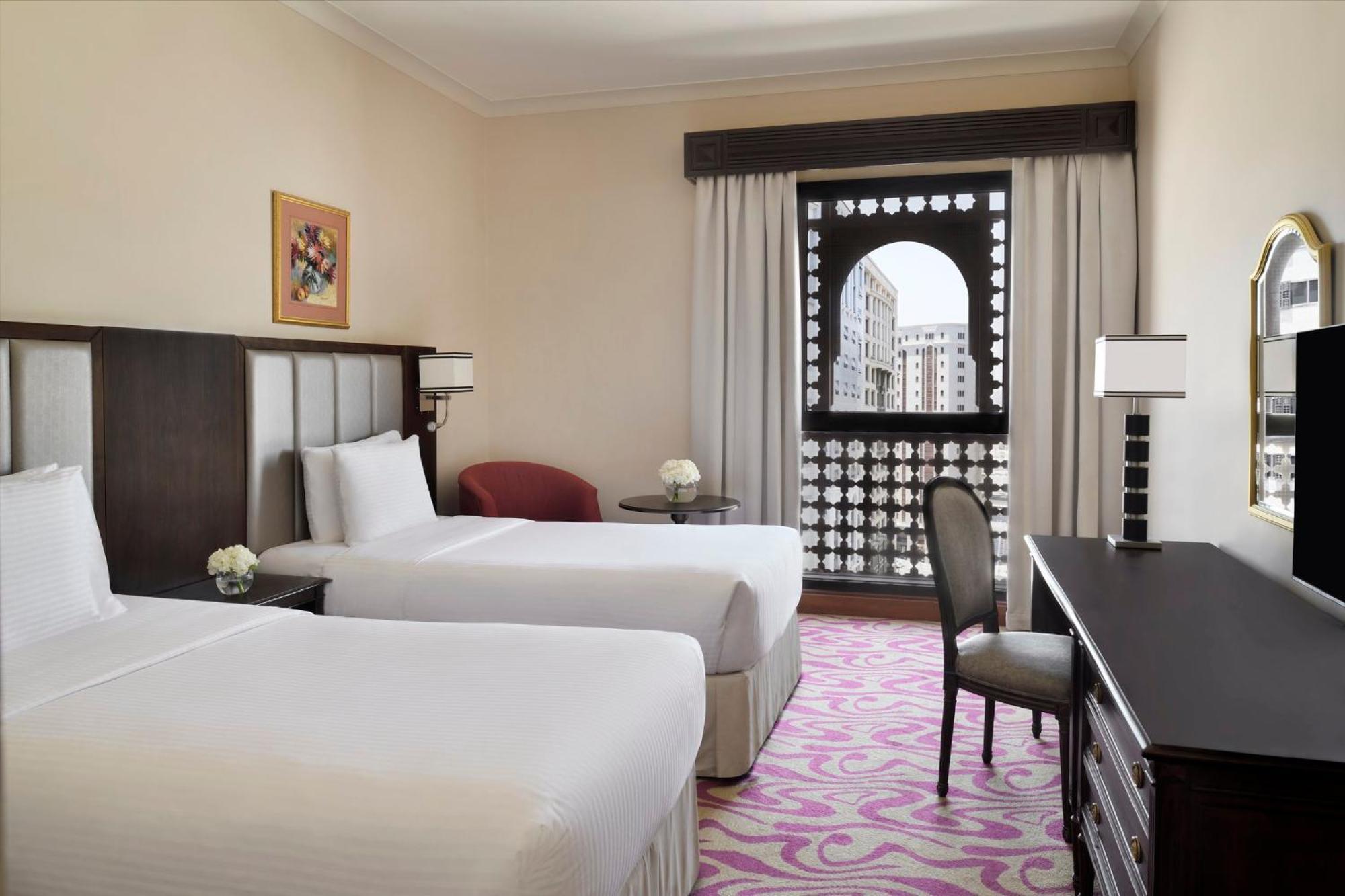 Hotel Intercontinental Dar Al Iman Madinah By Ihg Medína Exteriér fotografie The photo shows a hotel room featuring two single beds with white bedding. The room has a light, neutral color scheme and is decorated with minimal furnishings. There's a small seating area with a red chair and a round table, as well as a desk with a
