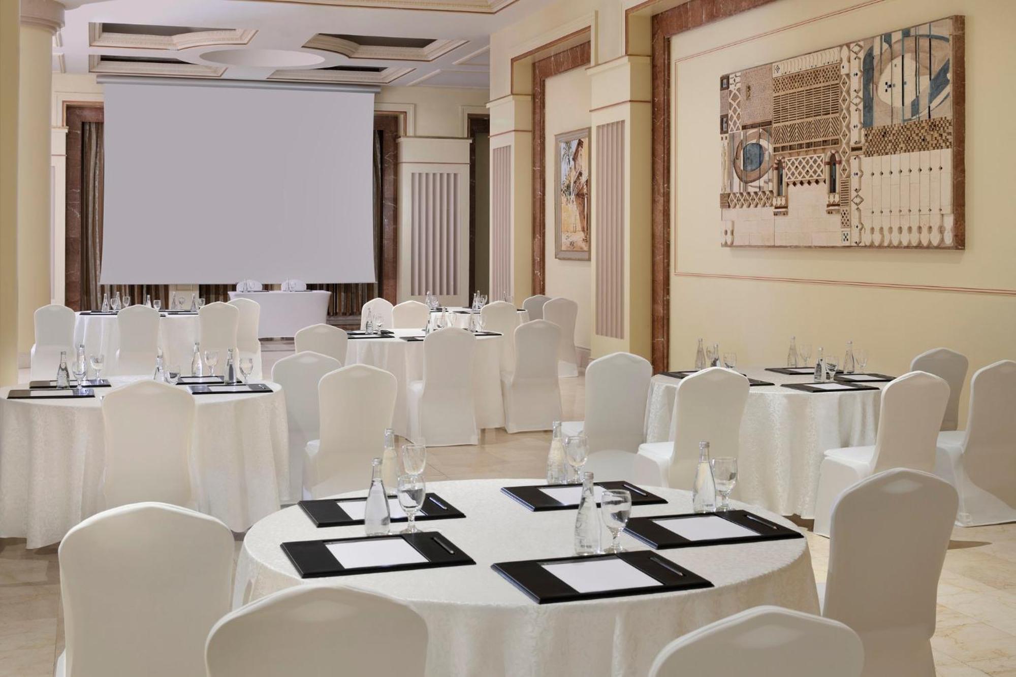 Hotel Intercontinental Dar Al Iman Madinah By Ihg Medína Exteriér fotografie The photo shows a well-decorated event space or meeting room. It features several round tables covered with white tablecloths and surrounded by white chairs. Each table has black placemats and glass water bottles, suggesting a formal setup for a gath