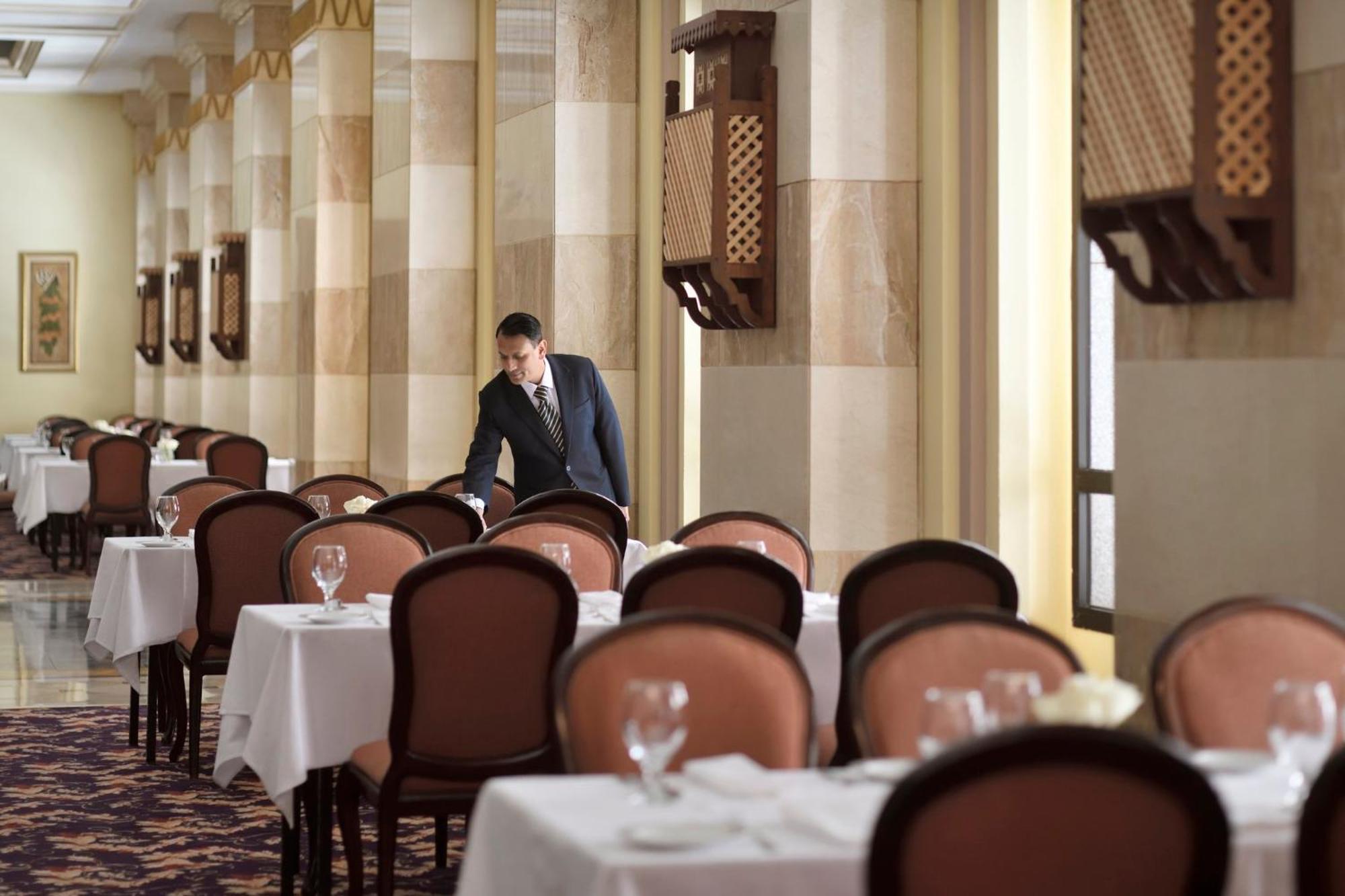 Hotel Intercontinental Dar Al Iman Madinah By Ihg Medína Exteriér fotografie The photo depicts a dining area in an elegant restaurant. There are several tables set with white tablecloths, each accompanied by chairs. A waiter, dressed in a formal suit, is seen arranging or tending to one of the tables. The restaurant features 