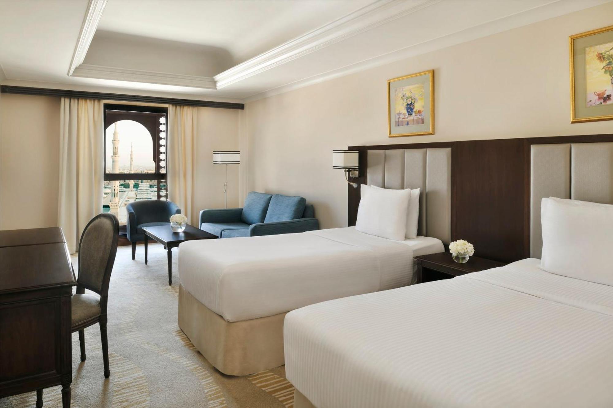 Hotel Intercontinental Dar Al Iman Madinah By Ihg Medína Exteriér fotografie The photo shows a well-appointed hotel room featuring two twin beds with white bedding. There is a small desk with a chair, a comfortable blue sofa, and a coffee table in the sitting area. The room has a warm, neutral color scheme, accented by two pa