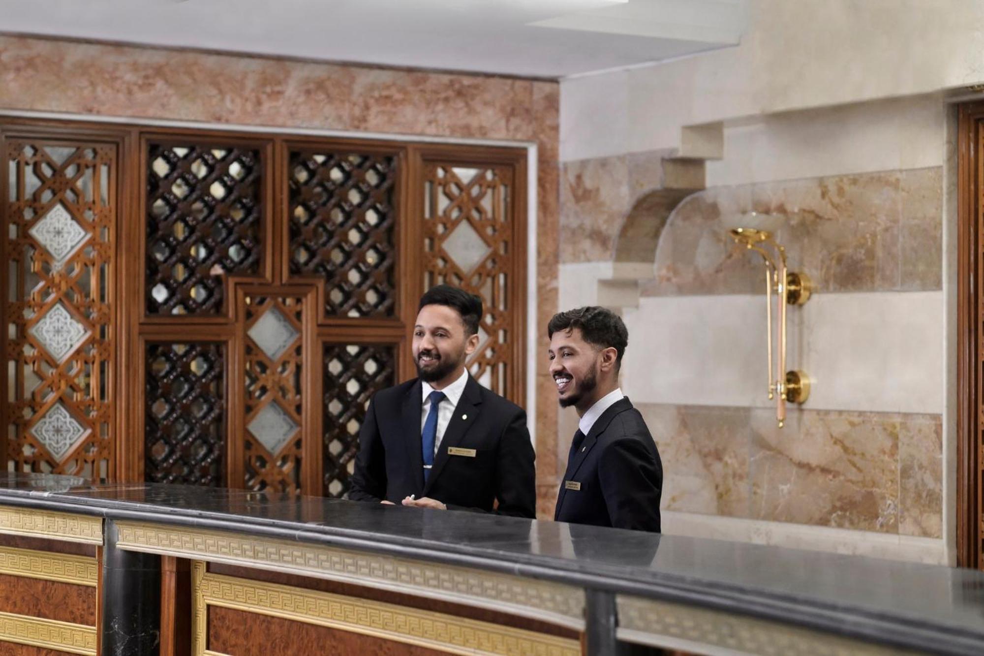 Hotel Intercontinental Dar Al Iman Madinah By Ihg Medína Exteriér fotografie The photo shows two men standing behind a reception desk in an elegant setting, likely a hotel or similar establishment. They are both dressed in formal attire, wearing suits and ties, and appear to be smiling or engaging in conversation. The backgro