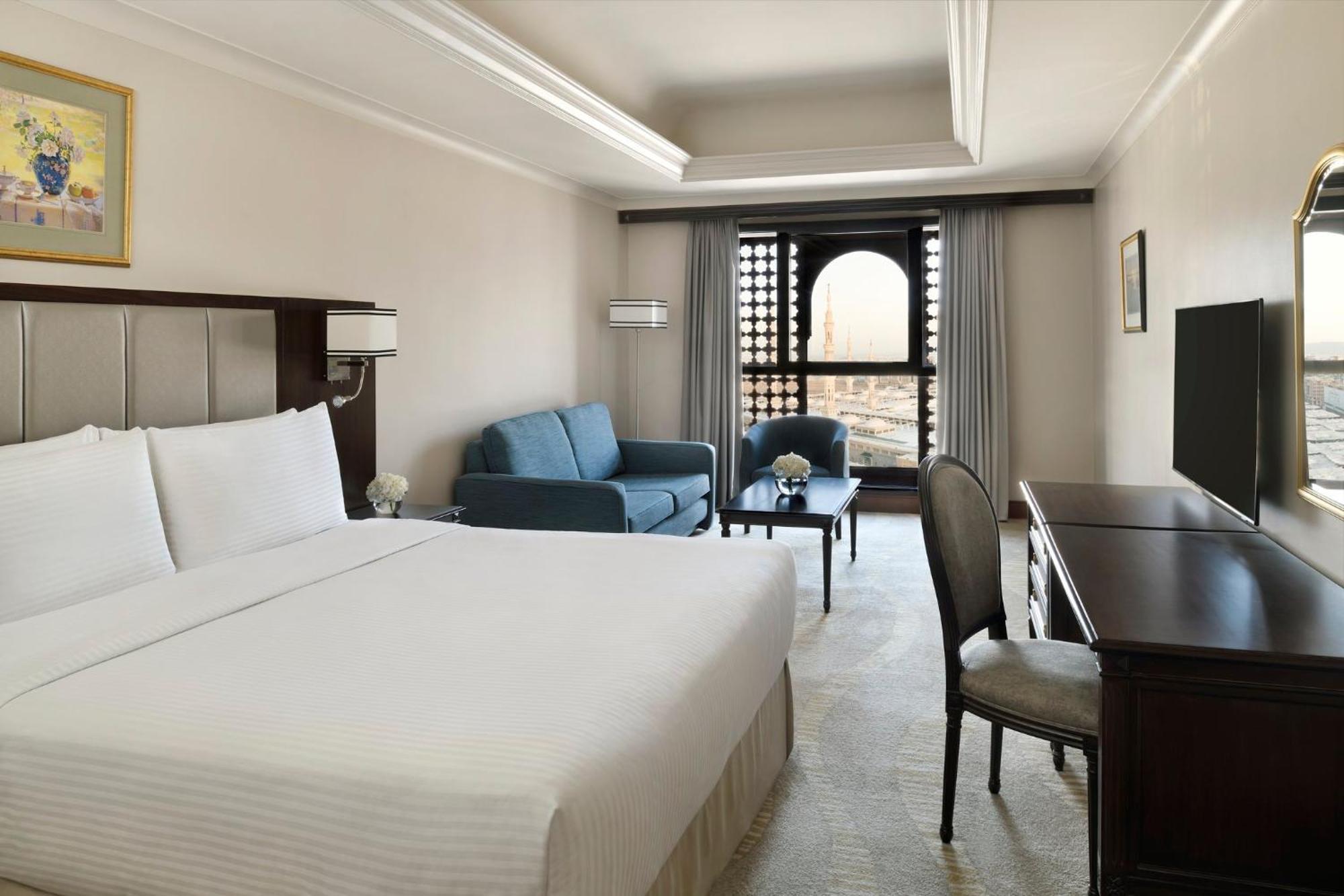 Hotel Intercontinental Dar Al Iman Madinah By Ihg Medína Exteriér fotografie The photo shows a hotel room designed with a modern and comfortable aesthetic. It features a large bed with white linens, a seating area with a blue sofa, and a small table. There is a wooden desk with a chair and a television opposite the bed. Natur