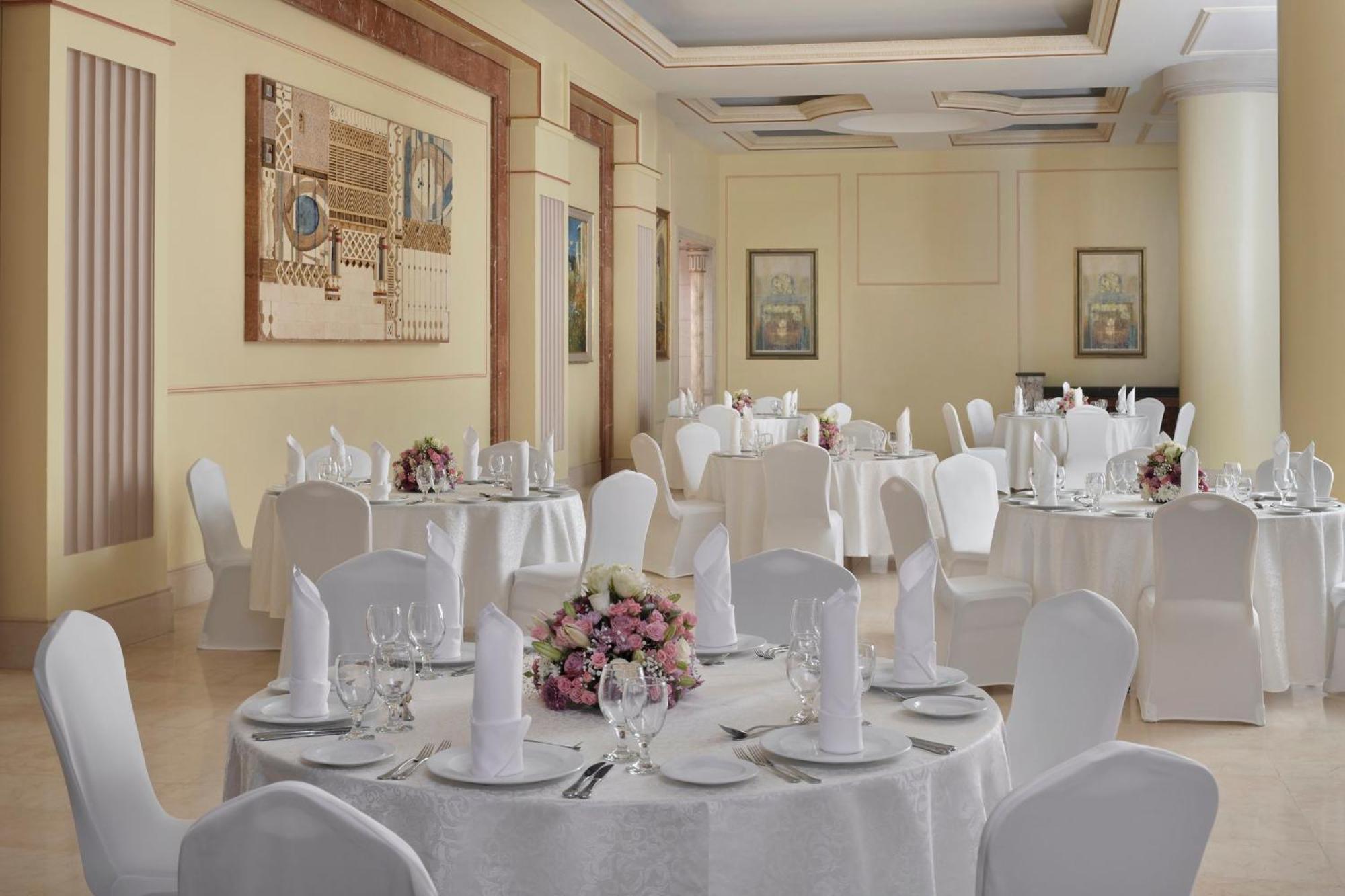 Hotel Intercontinental Dar Al Iman Madinah By Ihg Medína Exteriér fotografie The photo shows an elegantly arranged dining or function room. There are round tables covered with white tablecloths, each set with fine dining ware including glassware, utensils, and neatly folded white napkins. Some tables have floral centerpieces 