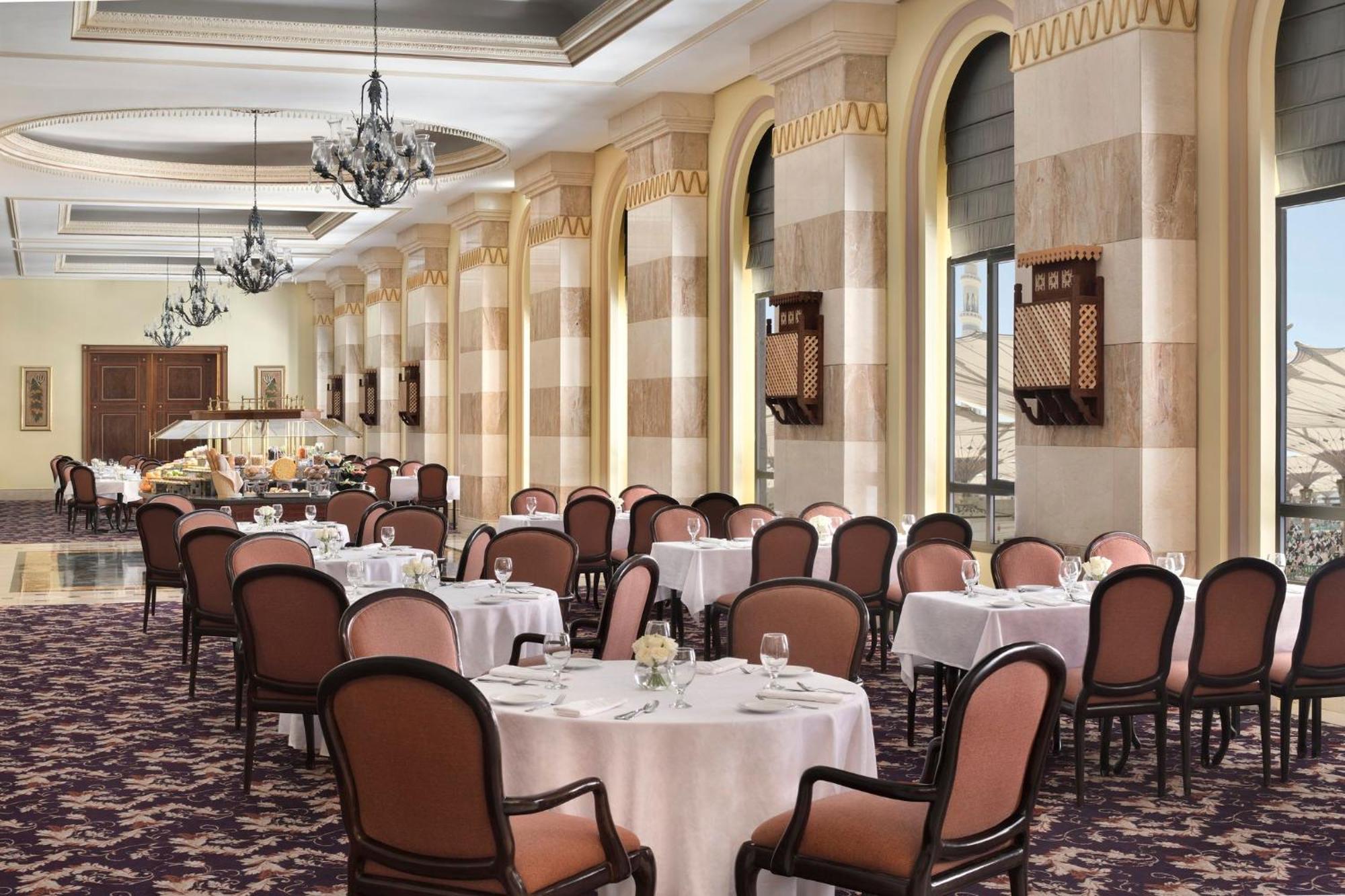 Hotel Intercontinental Dar Al Iman Madinah By Ihg Medína Exteriér fotografie The photo depicts an elegant dining room setting. It features several round tables covered with white tablecloths, each set with dishes and glassware. The room has a sophisticated ambiance, characterized by high ceilings adorned with chandeliers and 