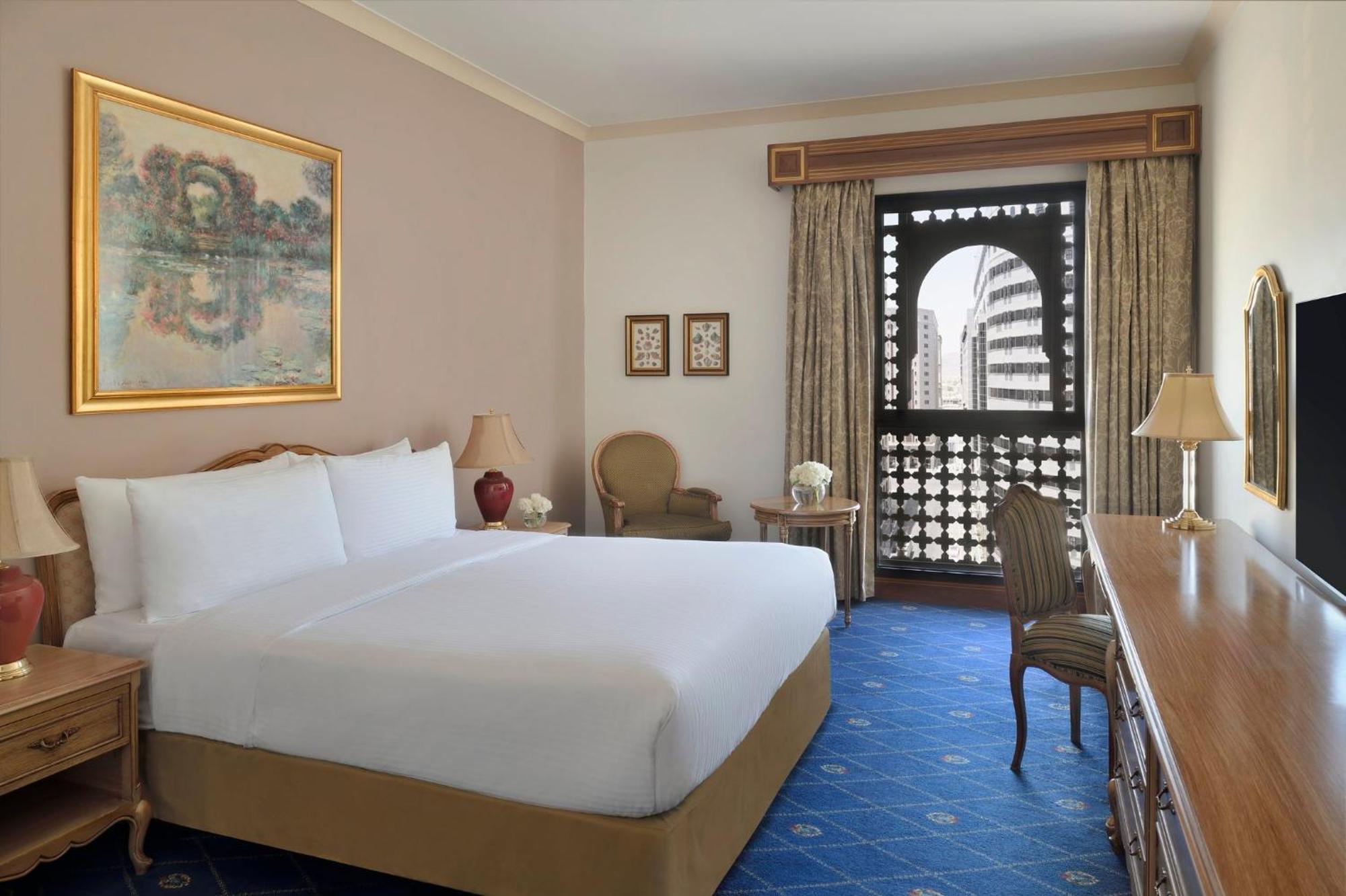 Hotel Intercontinental Dar Al Iman Madinah By Ihg Medína Exteriér fotografie The photo shows a hotel room that features a large, neatly made bed with white linens. There is a framed painting on the wall above the bed. To the side, a small seating area includes a chair and a side table. Opposite the bed, there is a wooden desk