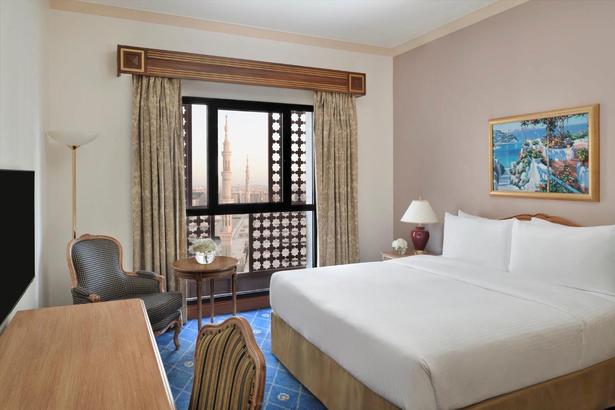 Hotel Intercontinental Dar Al Iman Madinah By Ihg Medína Exteriér fotografie The photo shows a hotel room with a modern design. There is a large bed with white linens placed against the wall. Next to the bed, there is a small bedside table with a lamp. A comfortable armchair is situated nearby, accompanied by a wooden desk. T