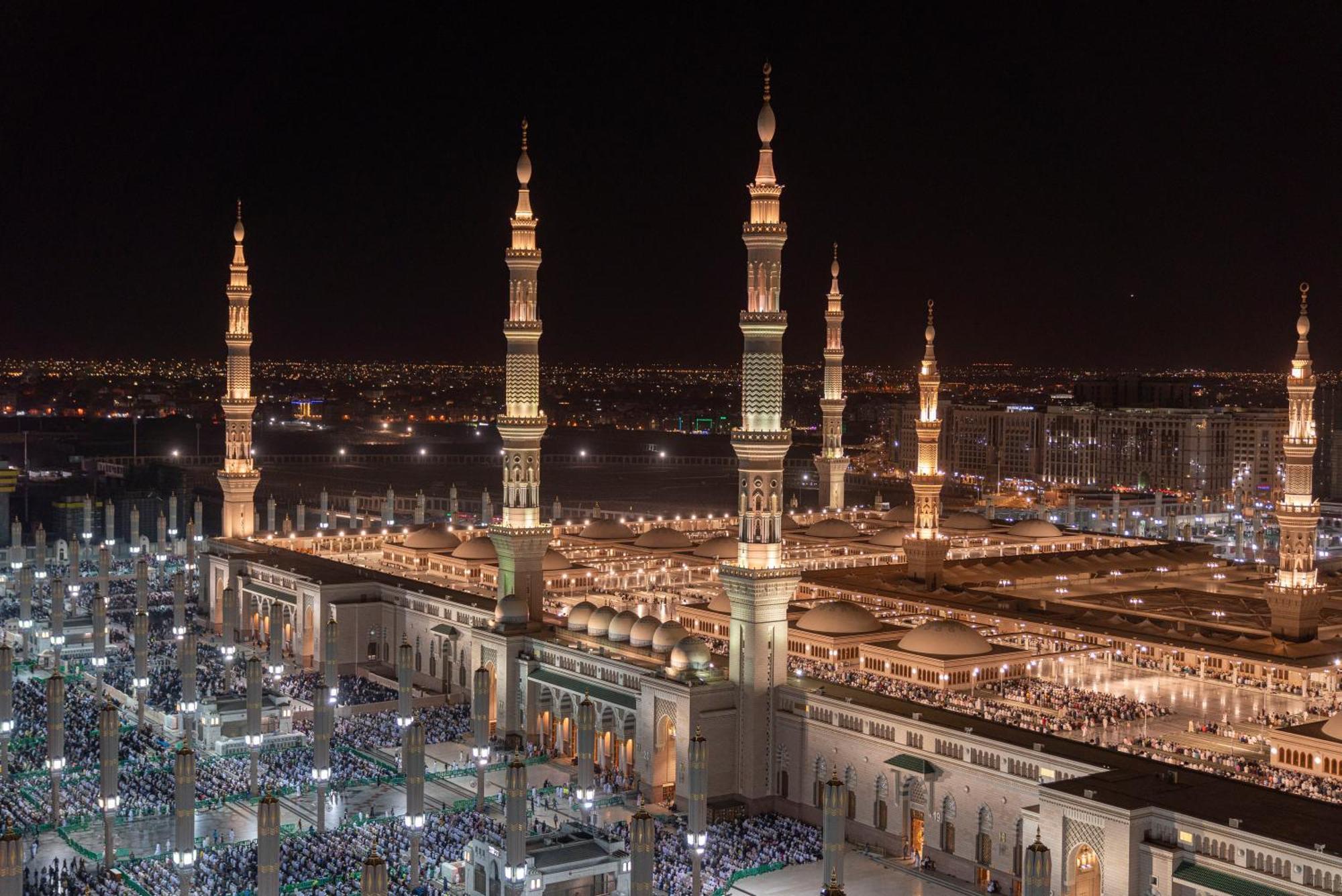 Hotel Intercontinental Dar Al Iman Madinah By Ihg Medína Exteriér fotografie The photo depicts a stunning nighttime view of a large mosque complex, prominently featuring tall minarets that are illuminated against the dark sky. The surrounding area is bustling with people, likely worshippers, gathered in the courtyard of the m