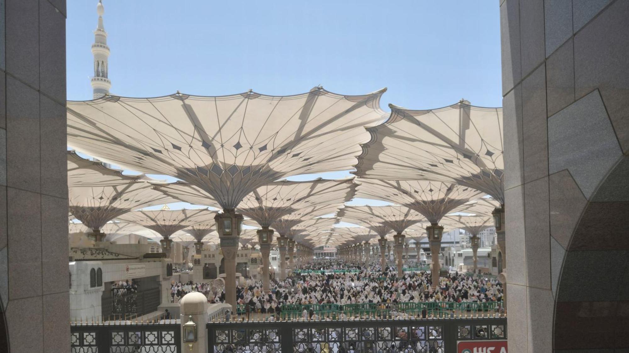 Hotel Intercontinental Dar Al Iman Madinah By Ihg Medína Exteriér fotografie The photo shows a large open area covered by expansive canopies, likely designed to provide shade for the people below. Underneath the canopies, there are many individuals gathered, possibly participating in a religious event or gathering. In the bac