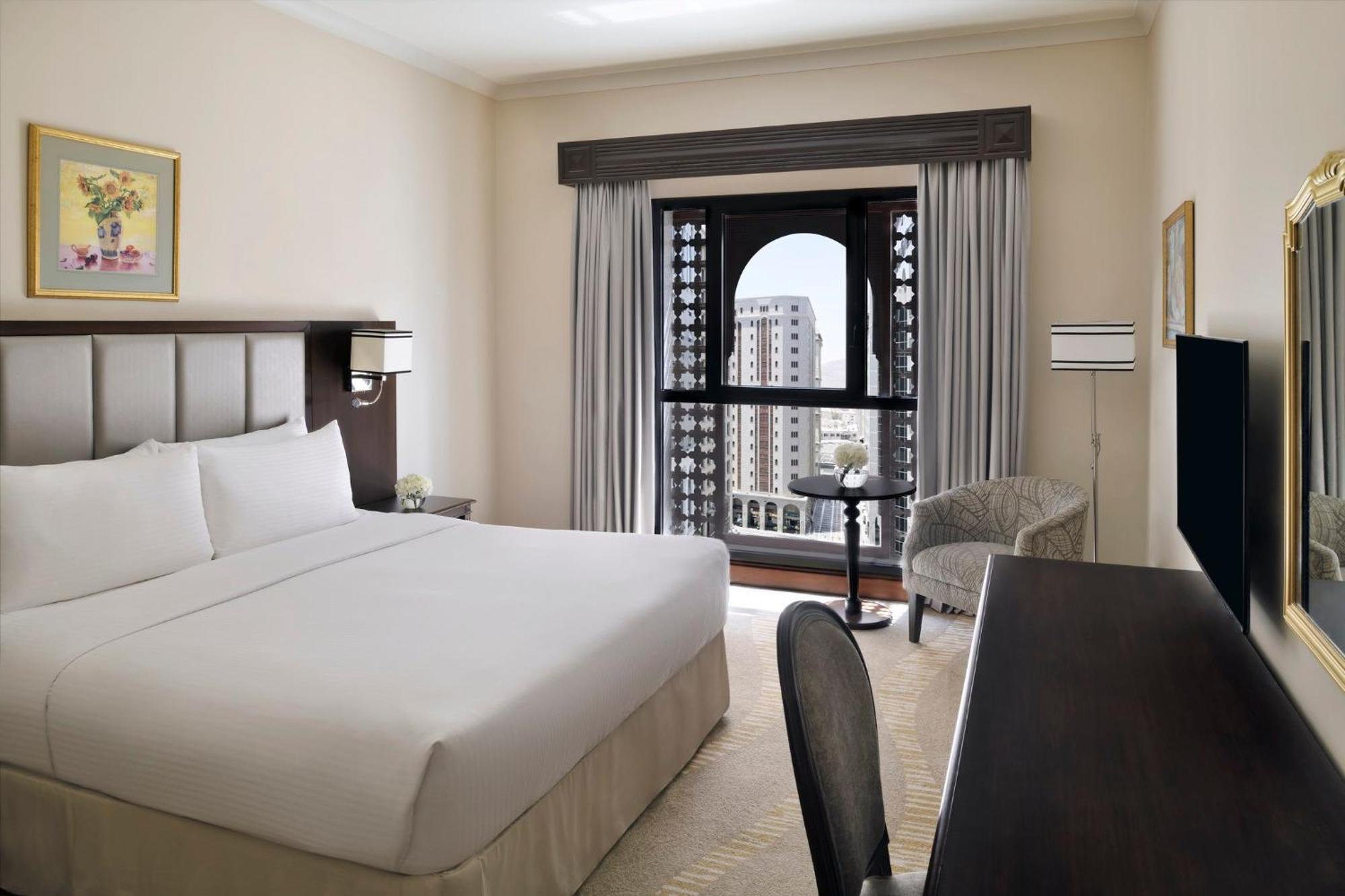 Hotel Intercontinental Dar Al Iman Madinah By Ihg Medína Exteriér fotografie The photo shows a hotel room with a modern and minimalistic design. It features a large bed with white linens, a side table, and a comfortable chair near a window. The window has a view of a cityscape, and elegant curtains frame the window. There is 