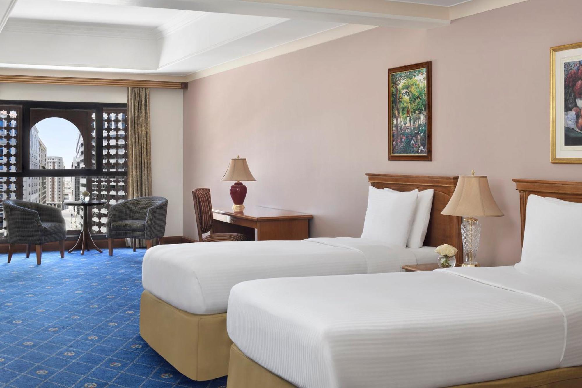 Hotel Intercontinental Dar Al Iman Madinah By Ihg Medína Exteriér fotografie The photo shows a nicely designed hotel room featuring two twin beds with white linens and beige bases. In the background, there's a wooden desk and a lamp, along with a pair of comfortable armchairs positioned near a large window. The window showcas