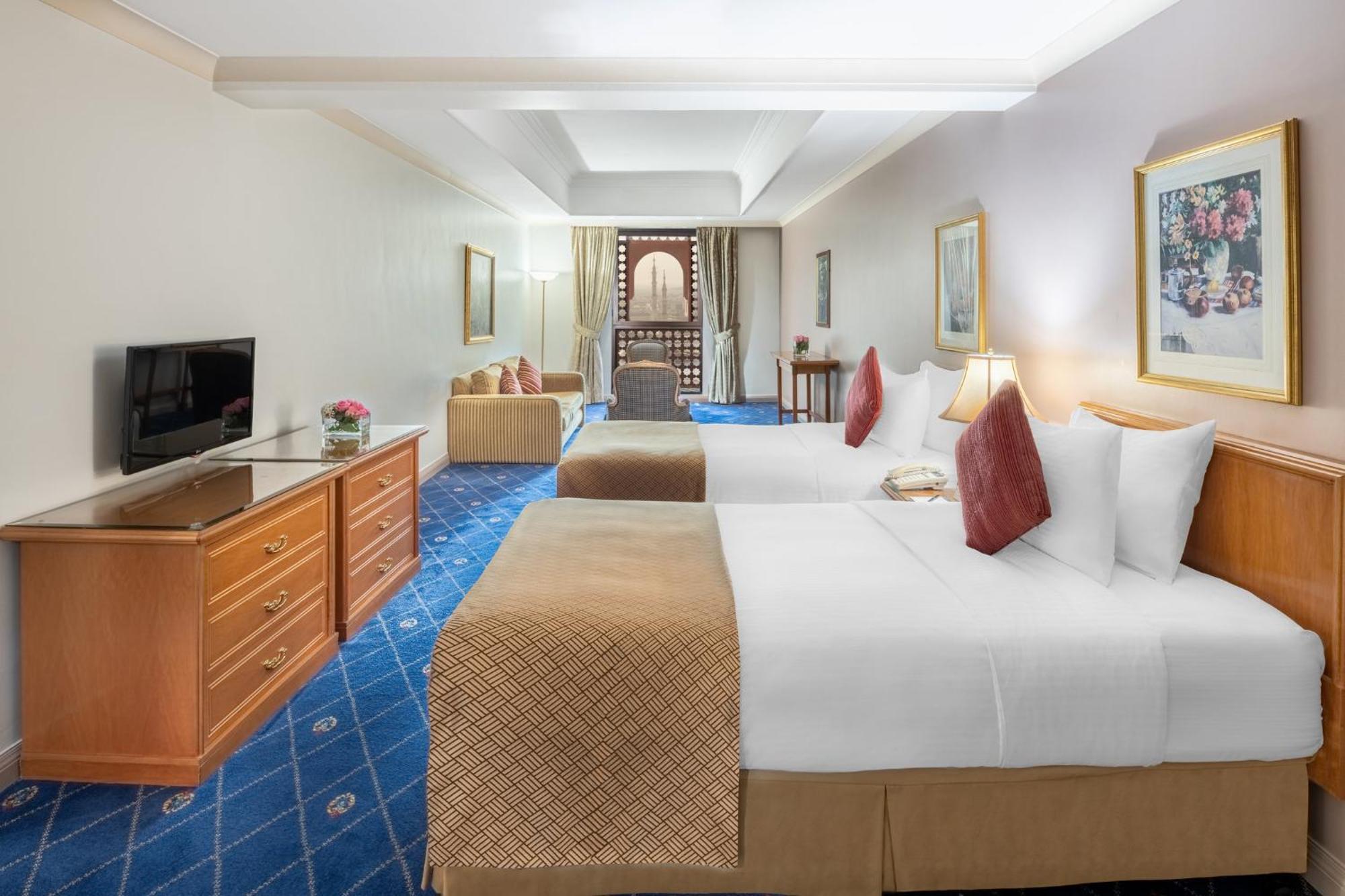 Hotel Intercontinental Dar Al Iman Madinah By Ihg Medína Exteriér fotografie The photo shows a hotel room featuring two beds with white linens and decorative pillows. There is a wooden dresser with a television on it. The flooring has a blue carpet with a pattern, and the walls are painted in a light color. The room is well-l