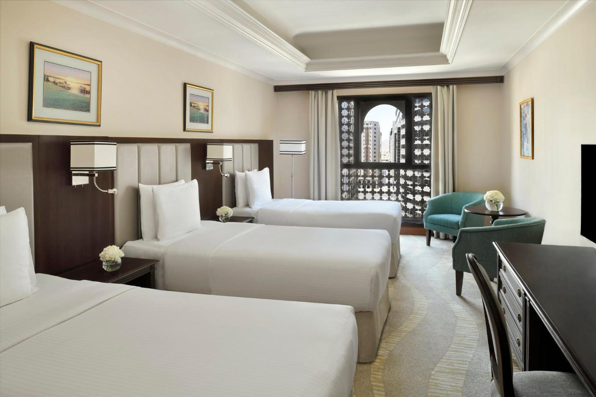 Hotel Intercontinental Dar Al Iman Madinah By Ihg Medína Exteriér fotografie The photo shows a hotel room with three single beds, dressed in white linens. There are two paintings on the walls, depicting serene landscapes. A large window allows natural light to enter, and it is adorned with elegant curtains. A smaller seating 