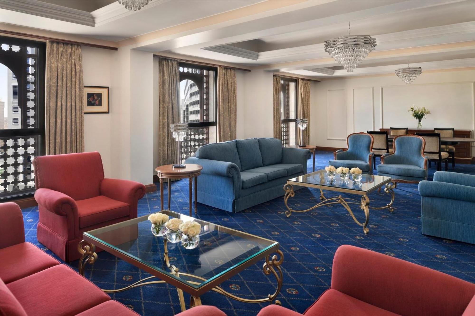 Hotel Intercontinental Dar Al Iman Madinah By Ihg Medína Exteriér fotografie The photo shows a luxurious living space or lounge area. It features a combination of furniture, including a blue sofa, several red armchairs, and glass-top coffee tables with gold accents. The flooring is carpeted in a deep blue, and the walls are a