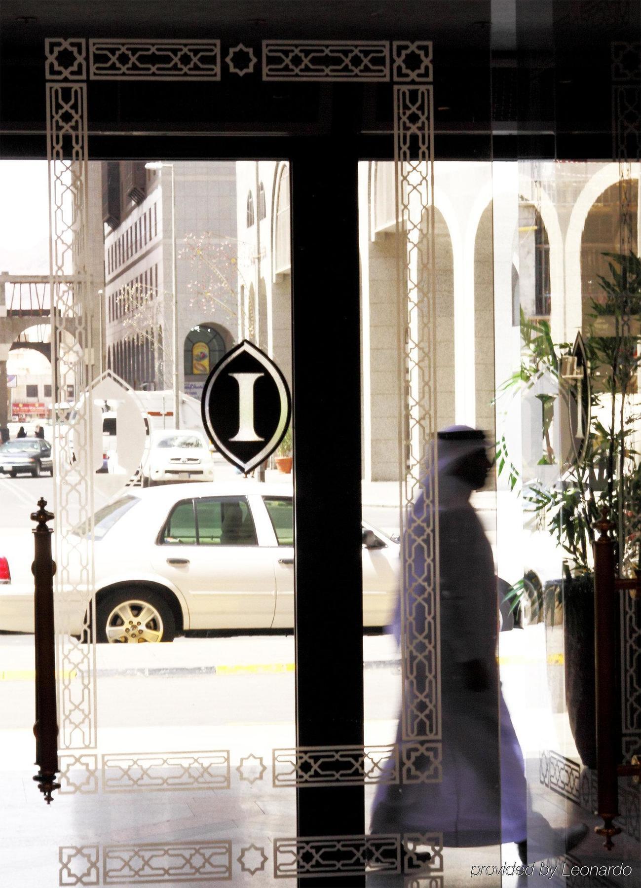 Hotel Intercontinental Dar Al Iman Madinah By Ihg Medína Interiér fotografie The photo shows a view through a glass door or window, featuring an elegant design with a prominent "I" symbol displayed. Through the glass, there are some parked vehicles visible, including a white car. In the foreground, a shadowy figure of a perso