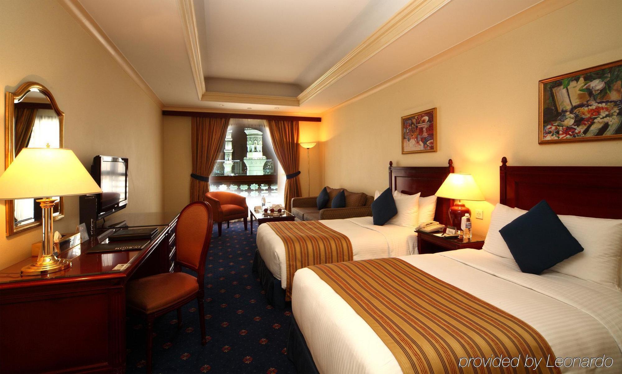 Hotel Intercontinental Dar Al Iman Madinah By Ihg Medína Pokoj fotografie The photo shows a hotel room featuring two beds with white linens and brown striped throws. There is a wooden desk and chair, alongside a television. The walls are painted in a warm tone, and there are decorative paintings. Large windows with curtain