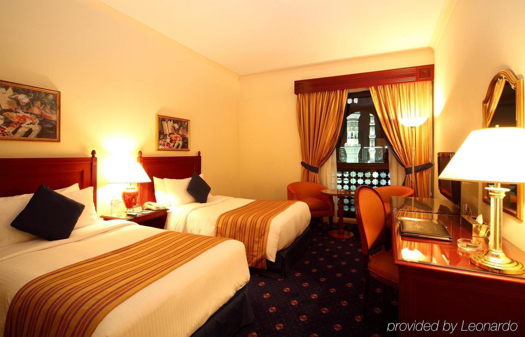 Hotel Intercontinental Dar Al Iman Madinah By Ihg Medína Pokoj fotografie The photo shows a hotel room featuring two twin beds with white linens and brown striped bedspreads. There are decorative pillows on the beds, and the room has warm-colored walls. To the side, there's a small sitting area with a round table and a cha