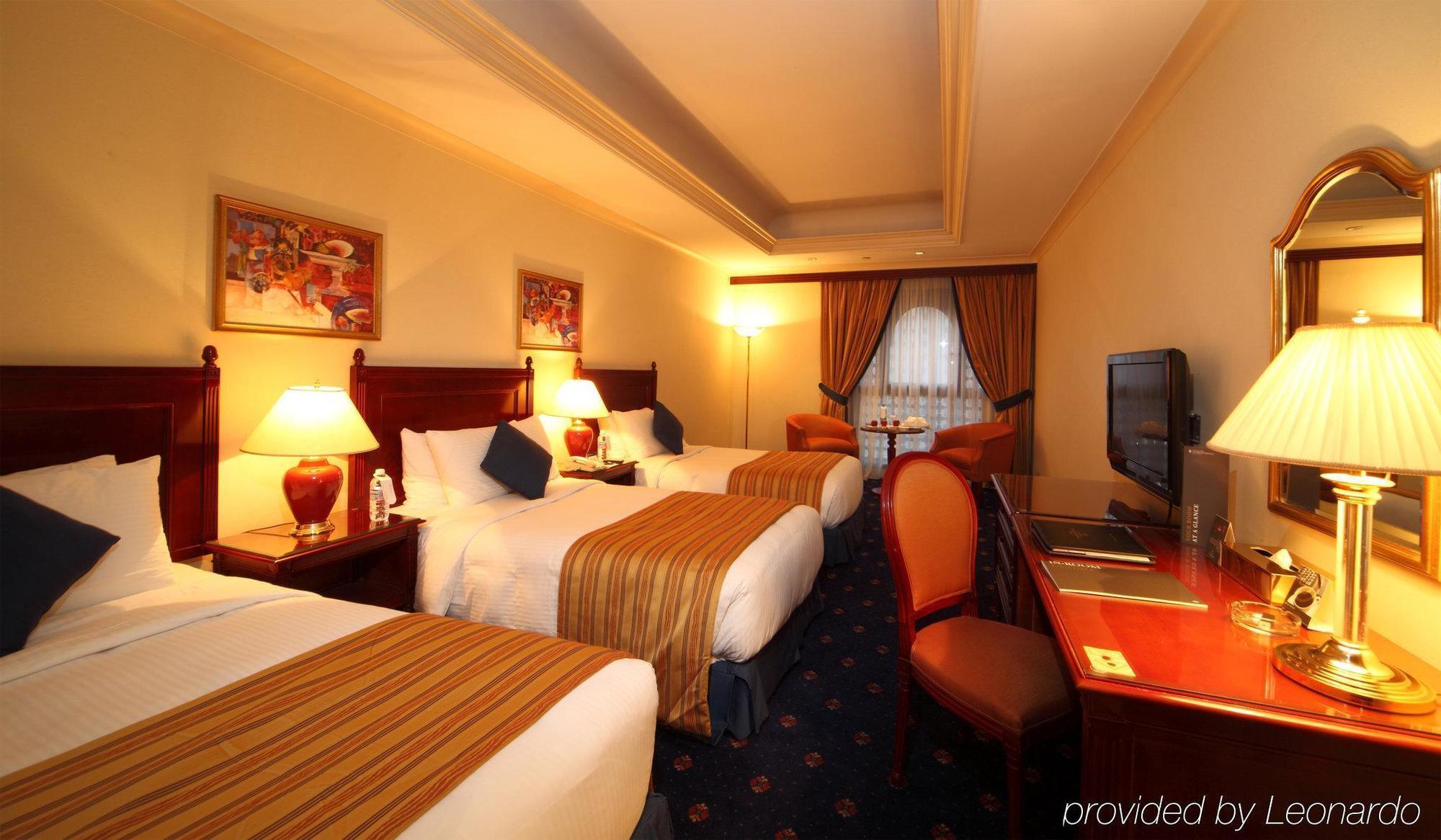 Hotel Intercontinental Dar Al Iman Madinah By Ihg Medína Pokoj fotografie The photo shows a hotel room with a warm and inviting atmosphere. There are two beds with white and gold bedding, complemented by decorative pillows. Each bed has a bedside lamp. In the background, there's a small seating area with a couple of chairs