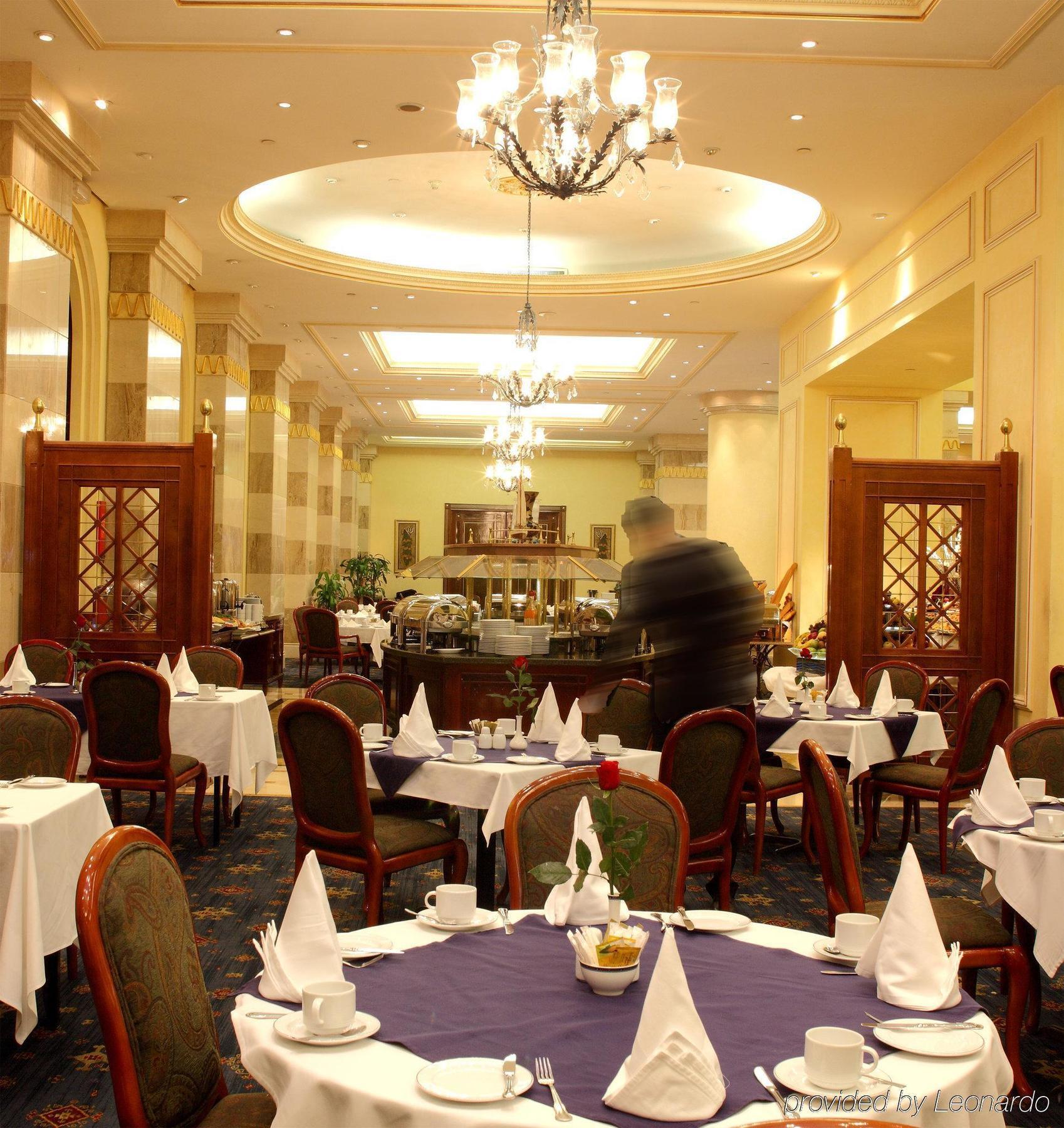 Hotel Intercontinental Dar Al Iman Madinah By Ihg Medína Restaurace fotografie The photo depicts an elegant restaurant interior. It features round tables covered with white tablecloths, each set with plates, cutlery, and folded napkins arranged into triangles. The space has a warm, inviting atmosphere with a mix of decorative e