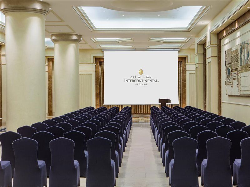 Hotel Intercontinental Dar Al Iman Madinah By Ihg Medína Exteriér fotografie The photo shows a conference or meeting room set up. There are rows of dark blue chairs arranged facing a large screen at the front of the room. The screen displays the logo of the Intercontinental hotel, indicating that this space is likely intended