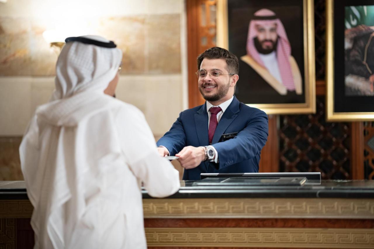 Hotel Intercontinental Dar Al Iman Madinah By Ihg Medína Exteriér fotografie The photo shows a hotel reception area. A male receptionist, dressed in a navy suit with a tie, is smiling and handing an item to a guest who is wearing traditional white attire, possibly a kandura or thobe. The background features elegantly decorate